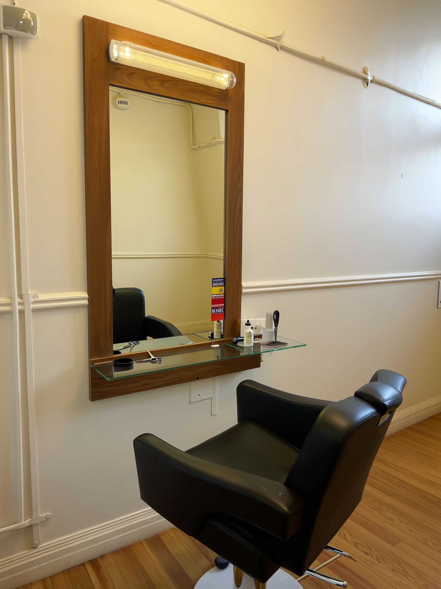 Een werkplek in de salon: een spiegel in een houten lijst met een lamp erboven, een glazen blad met schaar en flesjes, en een zwarte stoel ervoor.